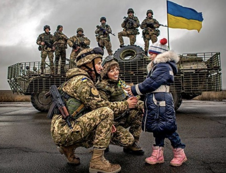 Foreground: a man and woman in military combat gear kneeling and talking to a child in a blue coat and pink boots. Background: Military personnel carrier bearing a Ukrainian flag alongside of which are six soldiers standing in a row, carrying weapons and wearing military combat gear.